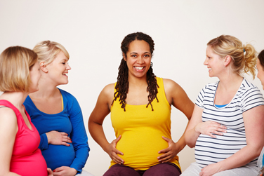 A group of pregnant women sit together, smiling and talking while resting their hands on their stomachs