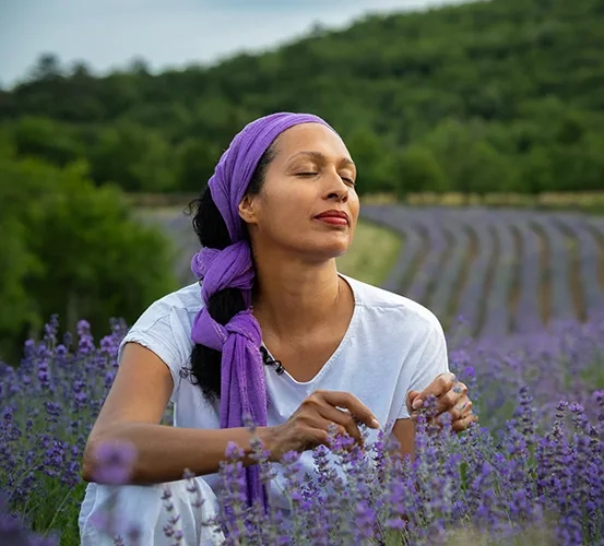 A woman smelling fresh lavender in a sunlit field, with rows of flowers stretching toward the horizon