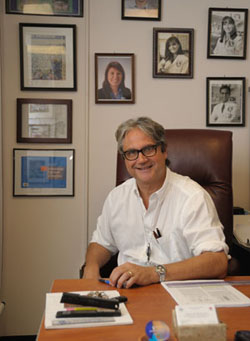 Dr. Francesco Marincola sitting at his desk in his office. On the wall behind him are framed photos of old fellows.