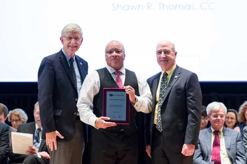 Dr. Francis Collins, Shawn Thomas and Dr. Jim Gilman stand on stage as Thomas holds a plaque