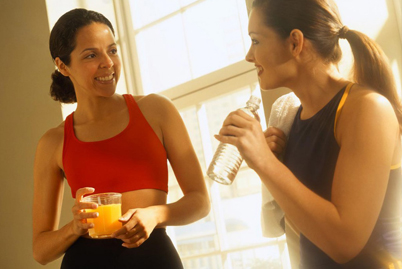 Picture of a woman drinking orange juice and smiling at another woman drinking water in workout clothes