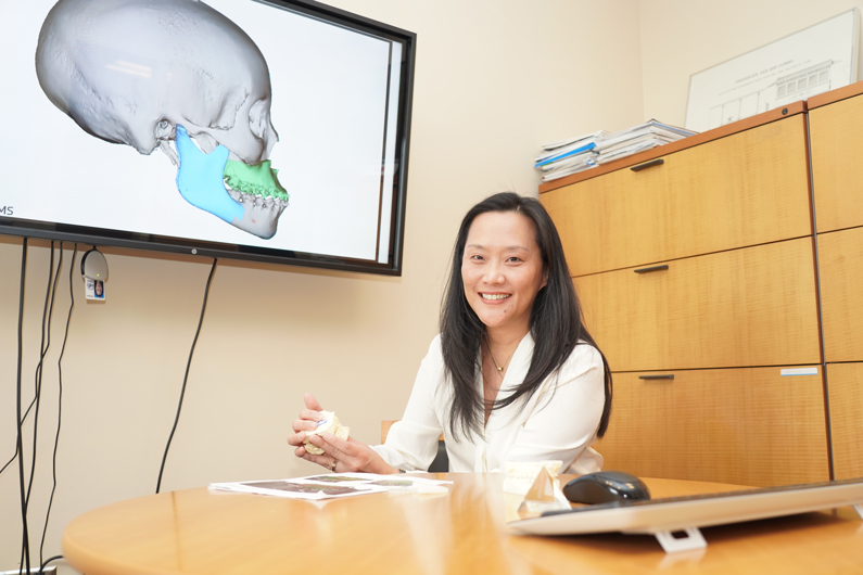 Doctor holds a stone model of a mouth, while the TV screen behind her has a 3-D model of a mouth
